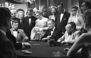 A black-and-white foto of a group of more than a dozen  expensively dressed white people clustered around a card table in a posh casino.