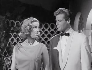 Honor Blackman and Roger Moore are standing outside in the garden at night. Both are formally dressed. They are looking at each other.