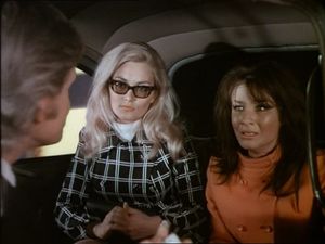 Three people are sitting in a small car: a blond man with his back to us, a blonde woman with dark glasses and a dark-haired woman, Kate O'Mara, who looks a bit dishevelled and distressed.