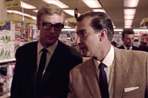 Michael Caine, wearing black-rimmed glasses and a suit,  is standing in a supemarket aisle with another man in a suit.
