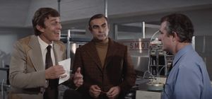 James Bond is in a lab, wearing a brown turtleneck. On each side of him is a man — the one on the left is excitedly showing a piece of paper to the one on the right.