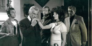 Five people are standing in an expensively decorated old room. From left to right: Tony Curtis in a vinyl jacket, an older man who is kissing a woman's hand, Roger Moore standing in the background, the woman, who is Joan Collins with a massive hairdo, and a middle-aged man with a moustache who is wearing a crumpled suit and a dark shirt.