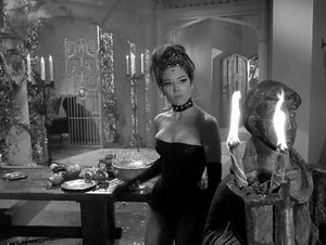 Emma Peel is standing in a Regency-styled room. There is a statue holding burning torches in the foreground.  She is wearing low-cut fetishwear and a spiked collar. She looks bored.