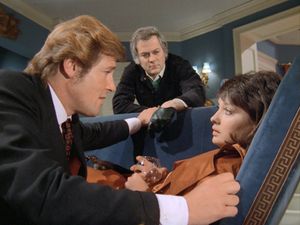 Roger Moore is crouched by a sofa, leaning over a young dark-haired woman who is lying on it with a glass of brandy in her hands.  Behind them, Tony Curtis looks on concerned.