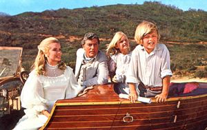 A young blonde woman in a white dress with a white hat, Dick Van Dyke in a white suit and a boy dressed in white are all sitting in a lovely wooden boat. There is a forested hill in the background.