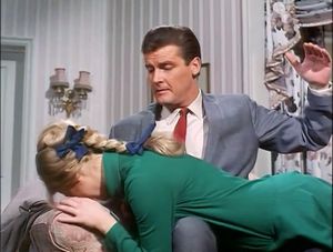 Roger Moore is sitting on a bed wearing a lovely suit. Over his knee is a young blonde woman covering her face. He has raised his hand as if to spank her.