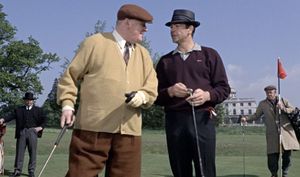 Auric Goldfinger and James Bond are standing on a golf course, each holding a golf club and staring each other in the face. Behind Goldfinger is Odd Job, a handsome Asian man in a top hat.