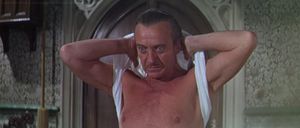 David Niven is standing in an expensively decorated old room. He is taking off his singlet in a determined-looking way.