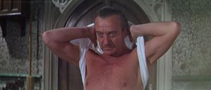 David Niven is standing in an expensively decorated old room. He is taking off his singlet in a determined-looking way.