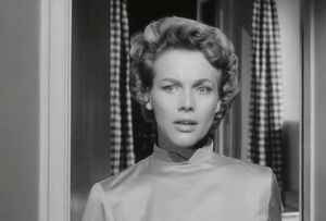 Honor Blackman, dressed as a housewife, is standing, looking anxious, in front of her curtained kitched window. 