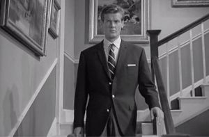A black and white photo of a very young Roger Moore wearing a lovely suit, descending the staircase of a suburban house.