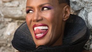 A close up of a black woman in strikingly coloured makeup with a high black collar; she has short hair and her tongue  is touching her top front teeth. It's May Day.