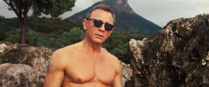 A shirtless Daniel Craig, wearing sunglasses, stands outside. There is a mountainous landscape in the background.