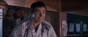 Sean Connery stands in a room with Japanese décor, wearing a Japanese dressing gown. He is wearing a wig which fails to make him look Japanese in any way.