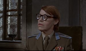 Rosa Klebb, a middle-aged woman with thick glasses and a military uniform, is sitting at a desk, looking sternly at something.