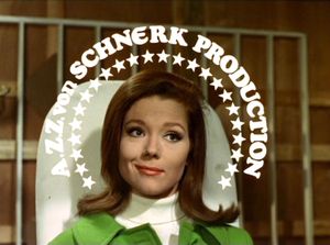 Emma Peel in a stylish green jacket is sitting in a chair smiling wryly to herself. Around her head, superimposed on the picture,  are the words 