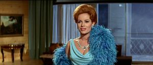 A beautiful red-haired woman is standing in a very 60s hotel room. She is wearing a blue dress with feathers around the collar.