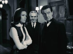 A black and white photo of Mrs Peel, Jean Le Mesurier and Steed standing urgently in a wood-panelled corridor. Le Mesurier stands between and slightly behind the others, with a devious look on his face.