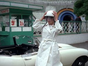 A young woman wearing a white suit is standing next to her expensive white sportscar and putting on a white hat. Behind her is an old-fashioned toolbooth.