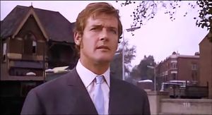 Roger Moore standing in a village street wearing a lovely suit with a pale blue tie. He looks very serious.