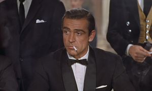 James Bond is sitting at a table in a casino, flanked by two men in suits. He has a cigarette hanging from his mouth.