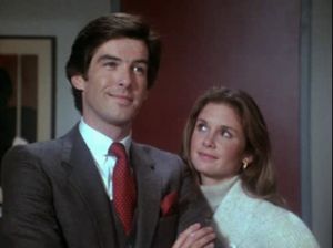 Pierce Brosnan is standing in an office in a lovely suit and red tie  smiling smugly. Stephanie Zimbalist is looking up at him admiringly.