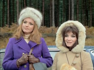 There are two young women in the woods, standing in front of a car: a tall blonde woman in a furry hat, and a short dark woman (Kate O'Mara) in a fur-lined hood.
