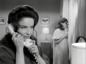 A black and white photo of two women in a bedroom. In the background, a woman in a nightdress is looking out the window with binoculars. In the foreground, a woman (Kate O'Mara) is talking conspiratorially  on the phone.