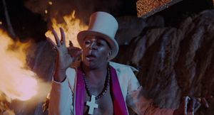 Standing in front of a bonfire is an African man, with half his face painted white. He is shirtless, but wearing a white top hat and a  white jacket.