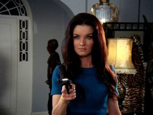 Kate O'Mara is standing in the living room of a suburban house, pointing a gun at someone. Her hair is even more massive than usual.